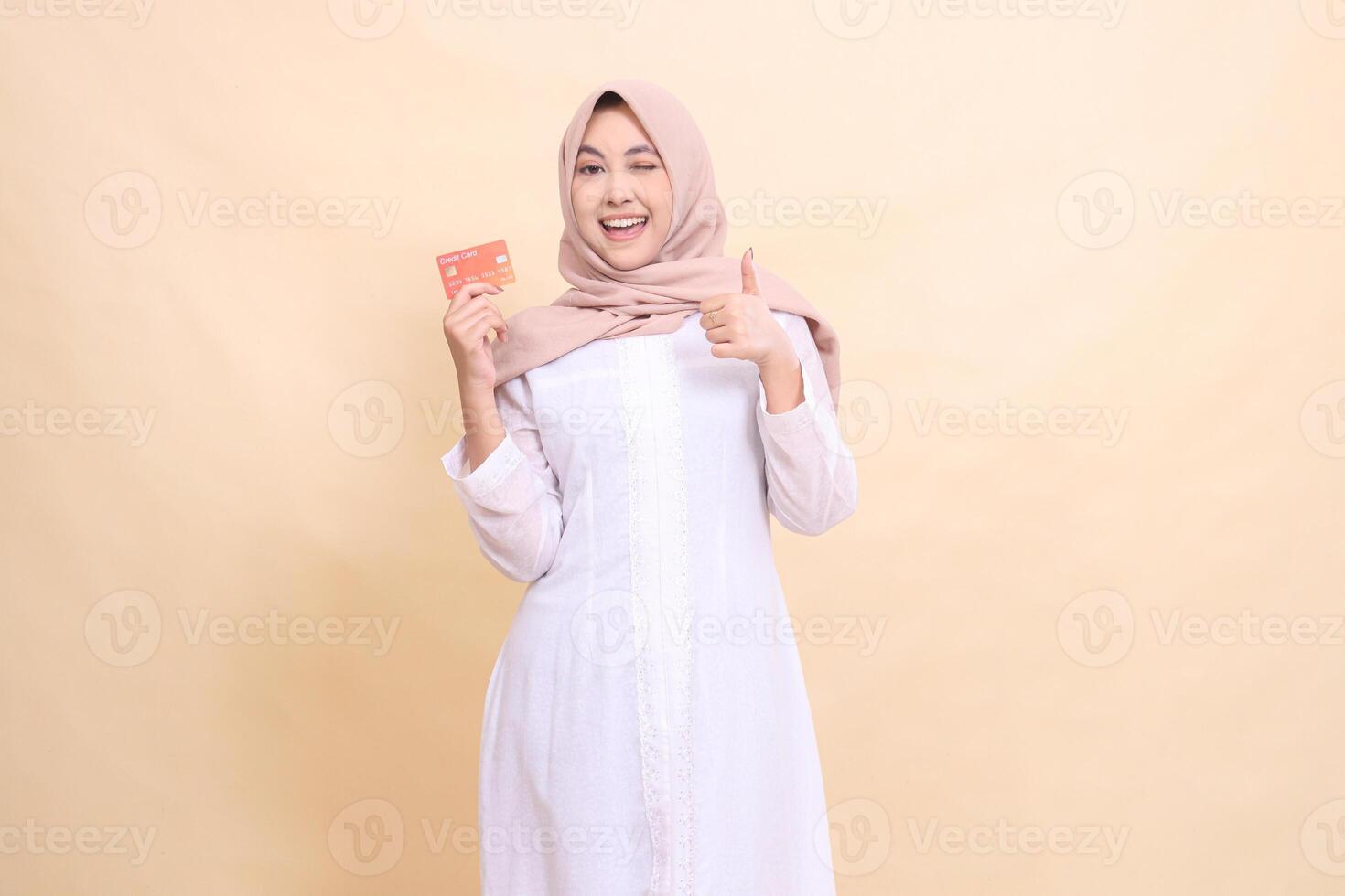 Asian Muslim woman in hijab smiling happily winking holding debit credit card in hand while giving thumbs up gesture good okay cool. Lifestyle, business and finance concept photo