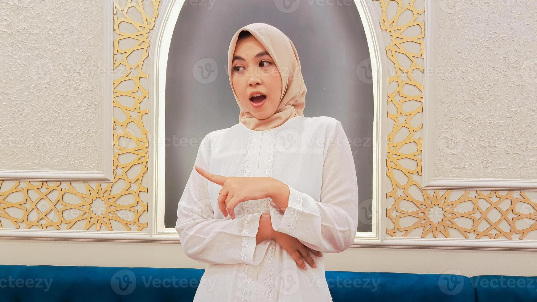 elegance of Asian woman wearing hijab candid expression of surprise, hand pointing to the right with Islamic ornamental calligraphy background photo