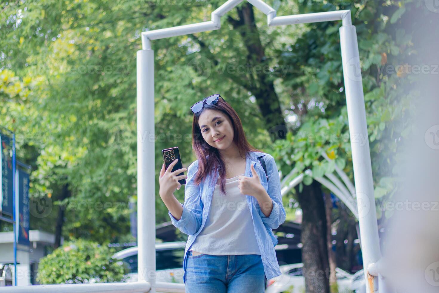 Beautiful smiling Asian woman using glasses holding smartphone gadget while giving good thumbs up gesture in campus park in sunny weather. female student for education, promotion and lifestyle concept photo