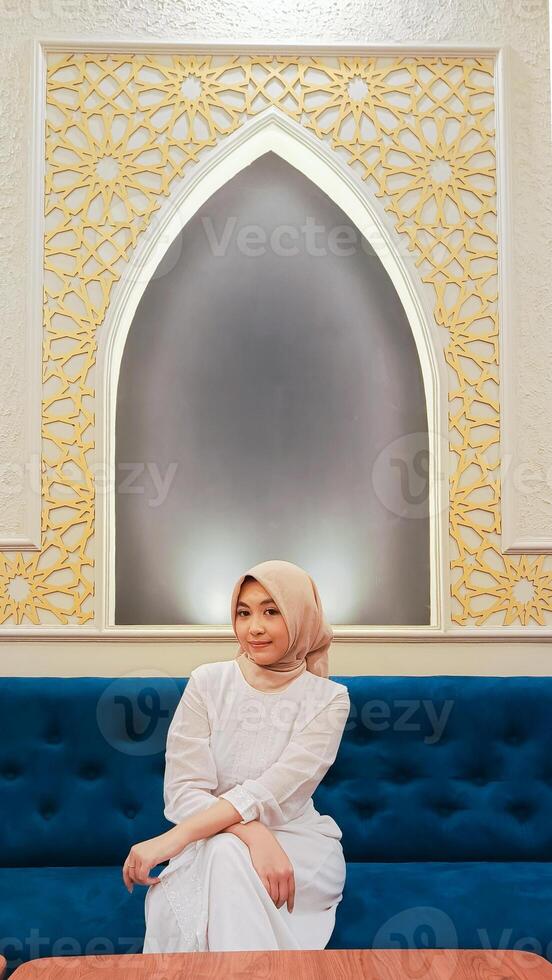 young woman indonesia forward smile expression hands crossed on thighs sitting on sofa with calligraphy background islamic mosque ornaments arabic nuances photo