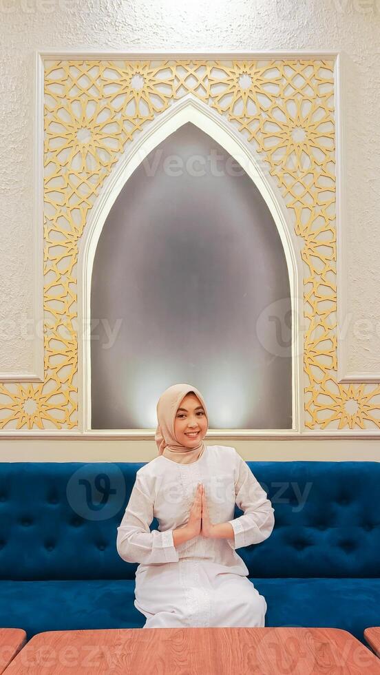 portrait of beautiful woman indonesia smiling cheerfully with hand gesturing welcome sawadikap sitting on sofa with calligraphy background of Islamic mosque ornaments in Arabic nuances photo