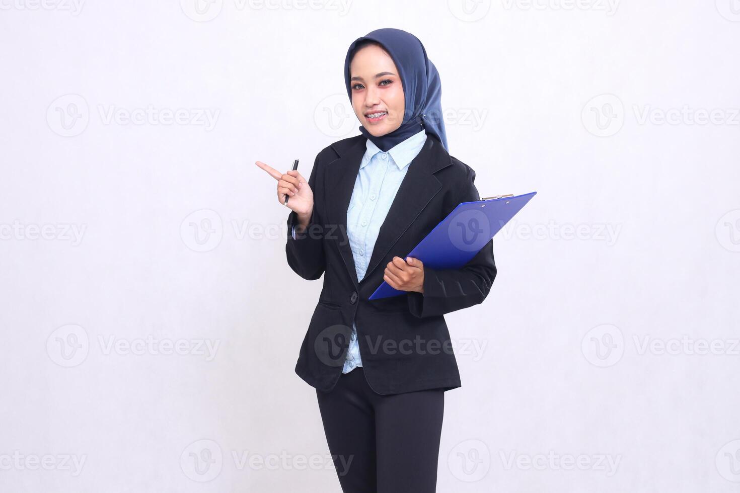Asian office woman wearing a hijab standing smiling cheerfully pointing right at the camera carrying a pen and hugging a clipboard. Beautiful Muslim women wearing blue shirts for business finance photo