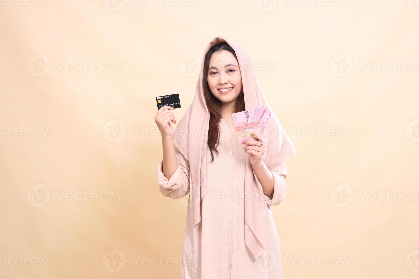 woman indonesia with a graceful smiling expression, holding her debit card up and carrying rupiah money. young beautiful for finance, advertising, fashion photo