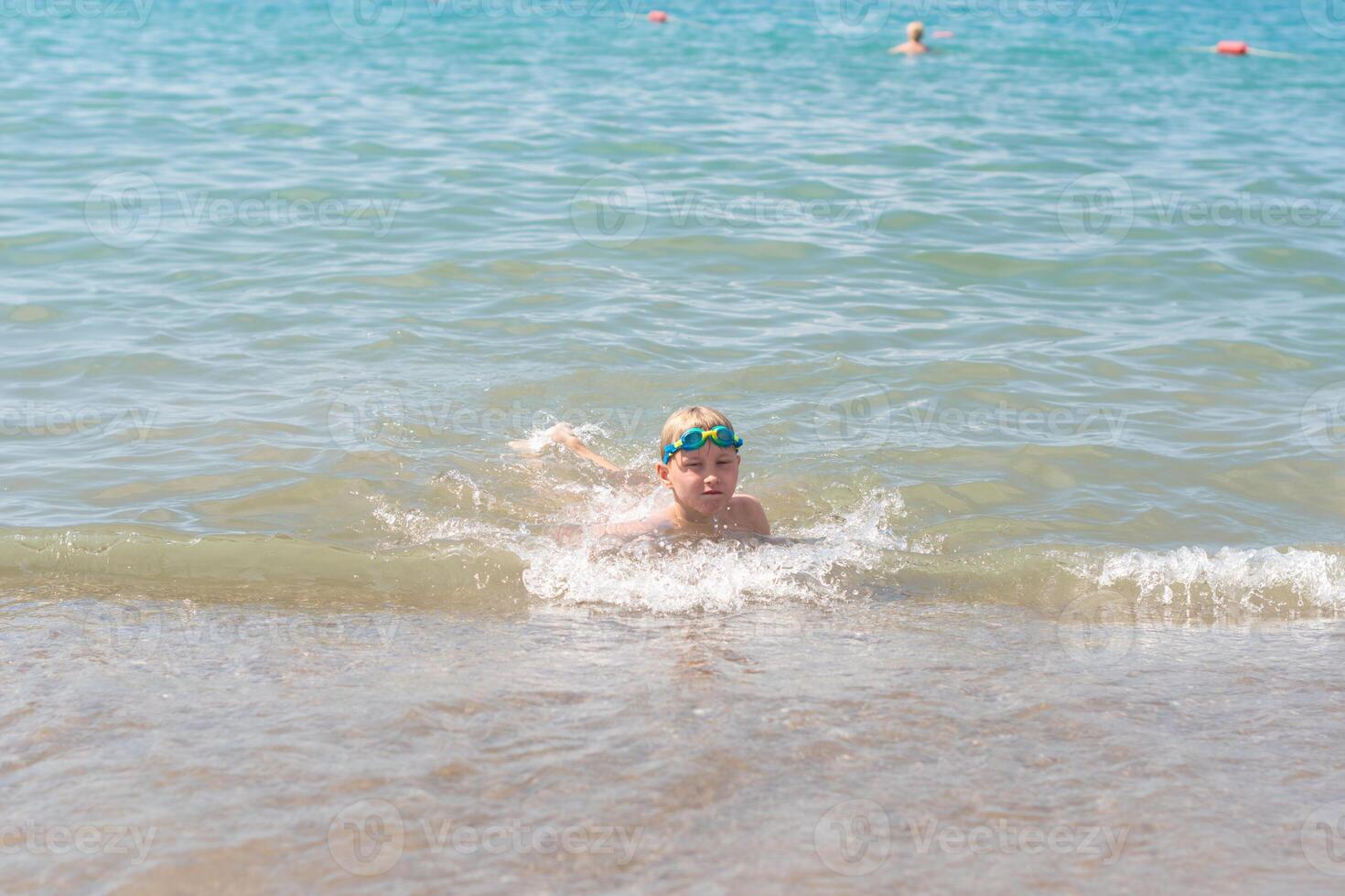 boy 8 years old in swimming goggles lies in the surf on the sandy beach of the blue sea, loves to swim during the summer holidays in Turkey, refreshing swim on a hot sunny day photo