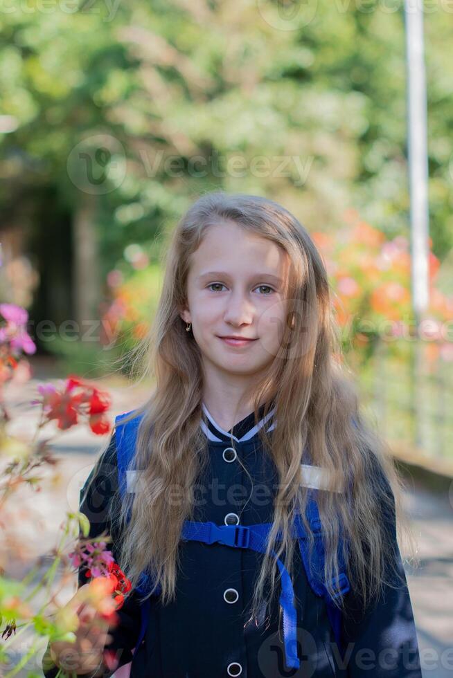 Back to school, portrait of a little girl from primary school outdoors, enjoying flowers in a flowerbed and looking at the camera with a smile, student dressed in dark blue uniform photo
