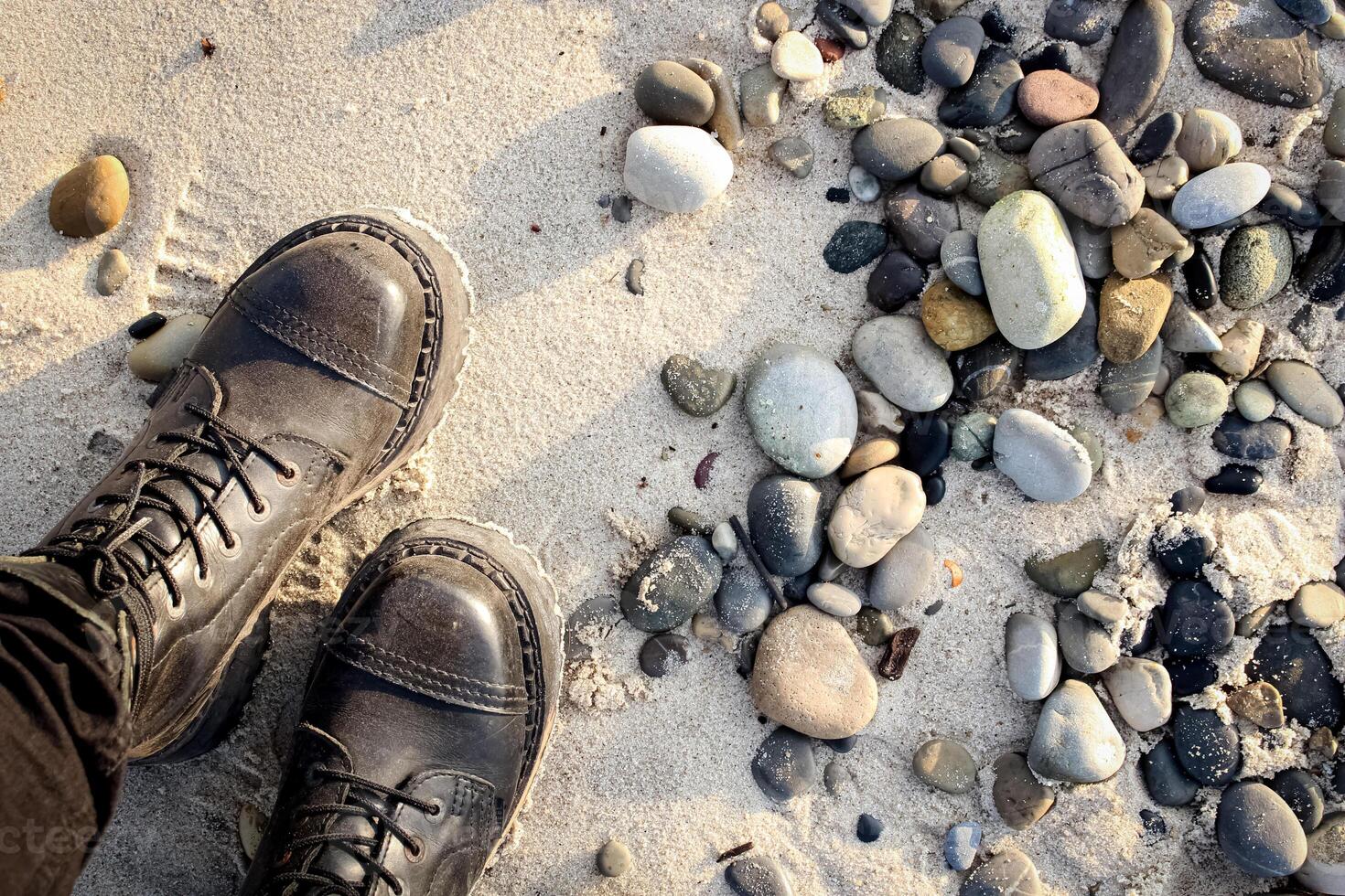 Legs of a standing female in rocker boots on beach sand with various rocks and pebbles in sunset light photo