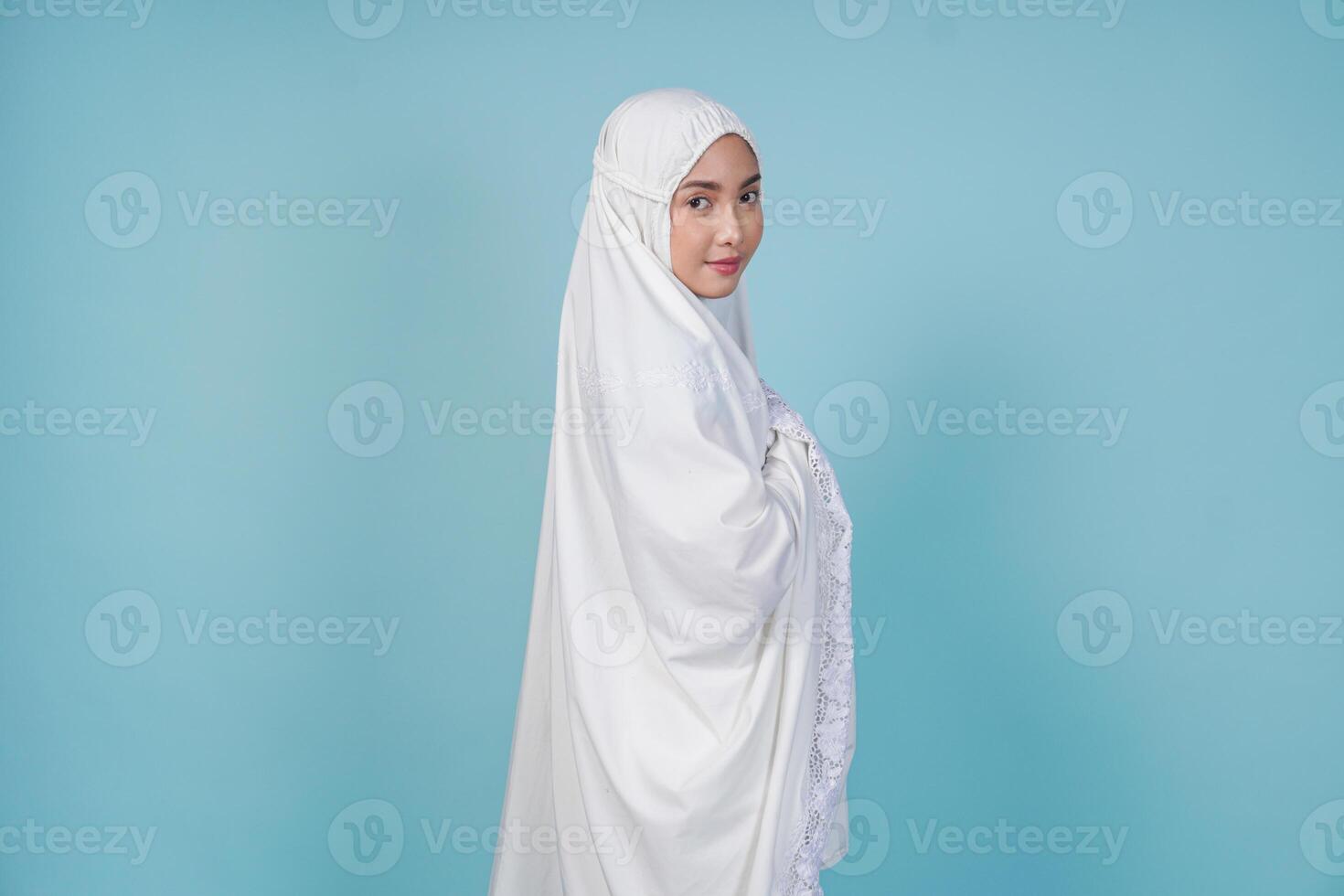Beautiful young Asian Muslim woman wearing prayer gown posing facing back, isolated by blue background. Hajj concept. photo