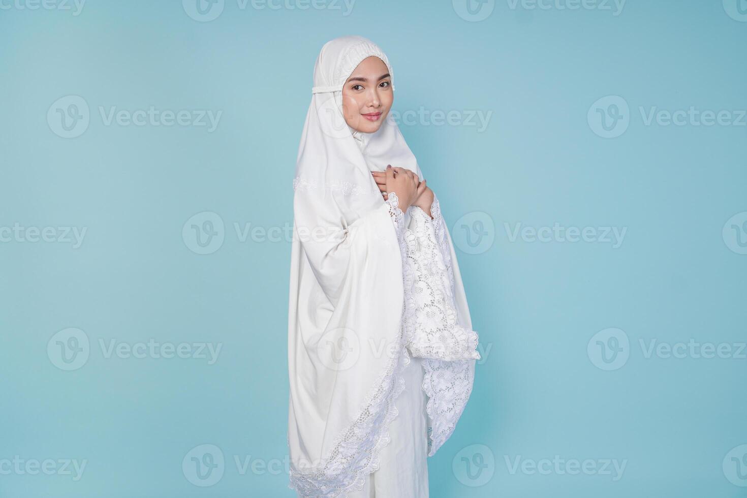Happy mindful thankful young Asian Muslim woman hand on chest wearing prayer gown over isolated blue background. Hajj concept. photo