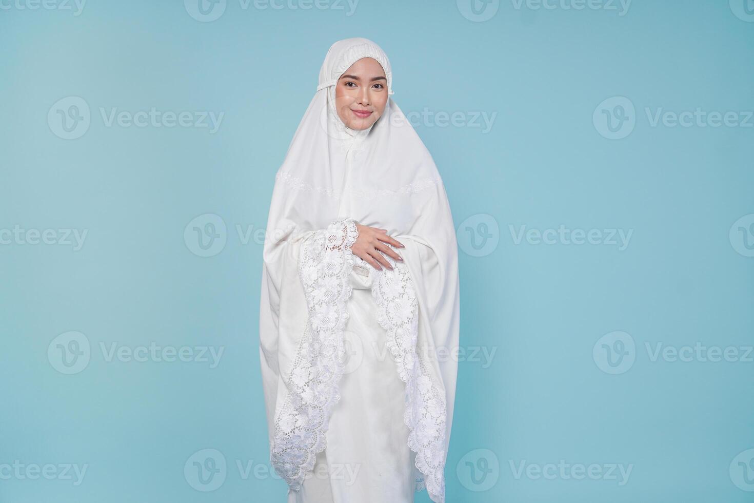 Young Asian Muslim woman wearing prayer gown smiling with hands folded gesture, isolated by blue background. Hajj concept. photo