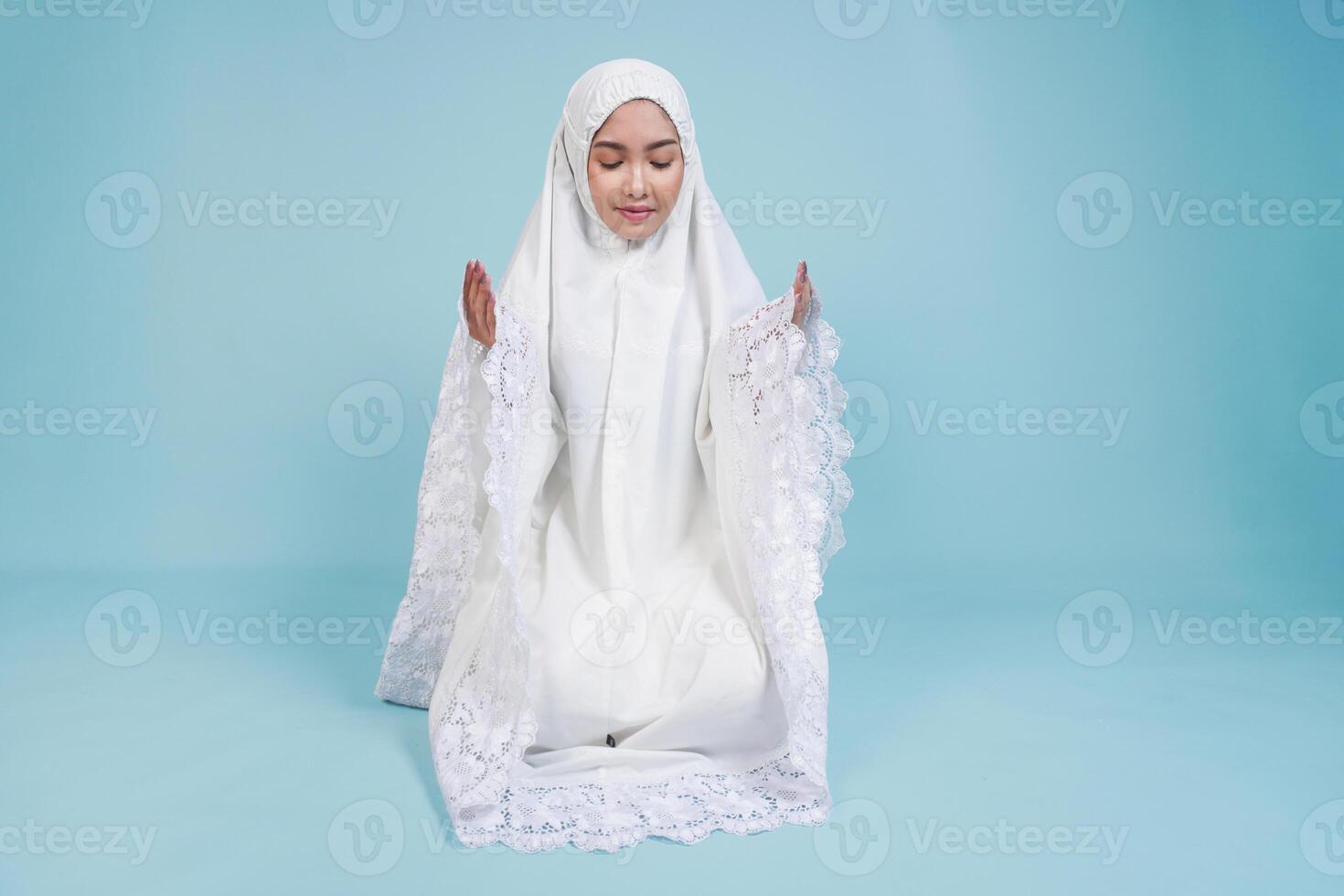 Young Asian Muslim woman in prayer gown sitting down and praying with closed eyes over isolated by blue background. Hajj concept. photo