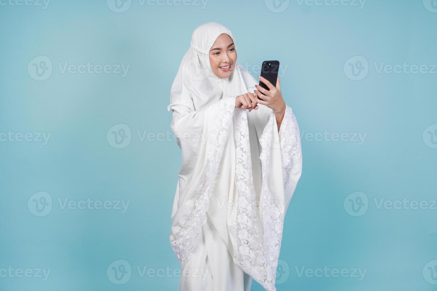 Attractive Asian Muslim woman in prayer gown using phone to text or browse internet, isolated by blue background. Hajj concept. photo