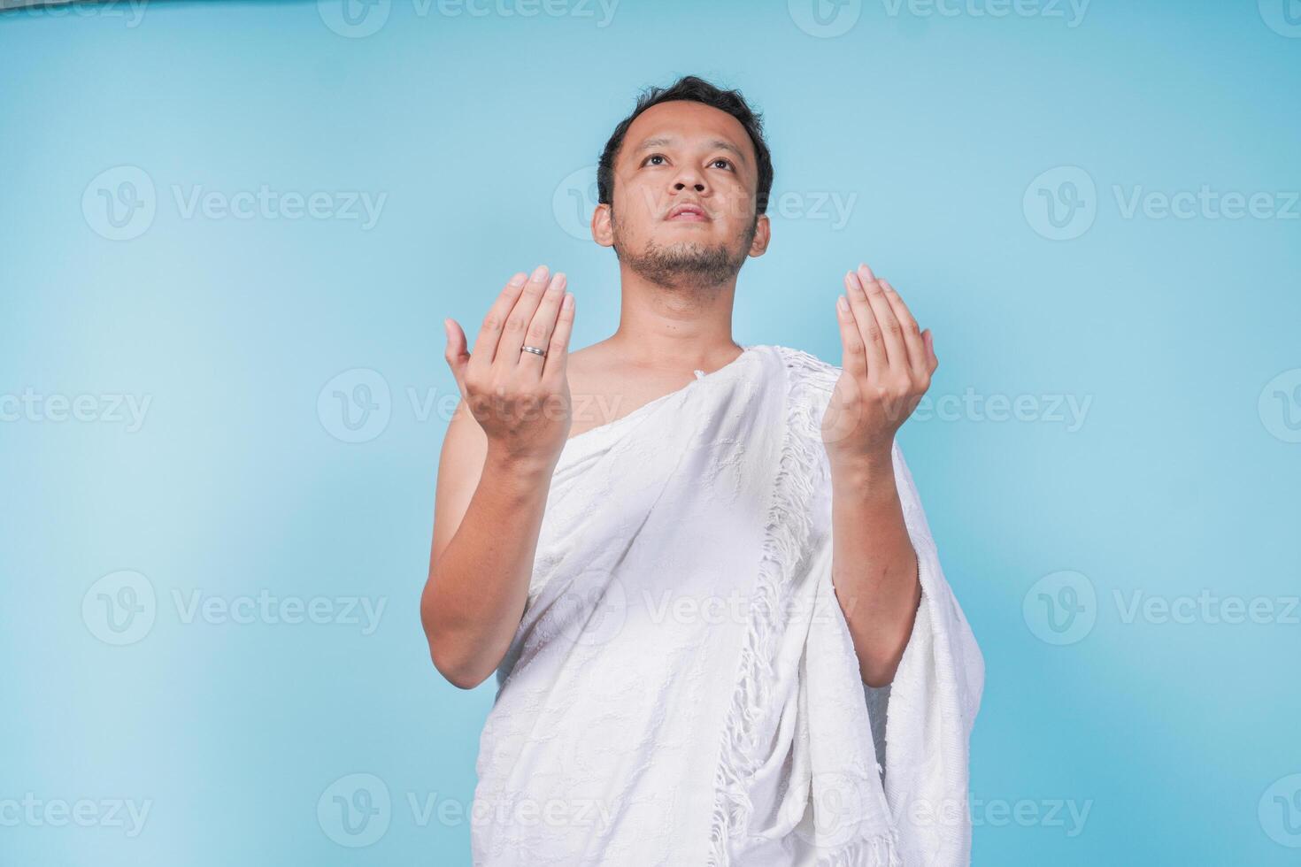 Asian Muslim man wearing ihram praying and lifting head up over isolated by blue background. Hajj concept. photo