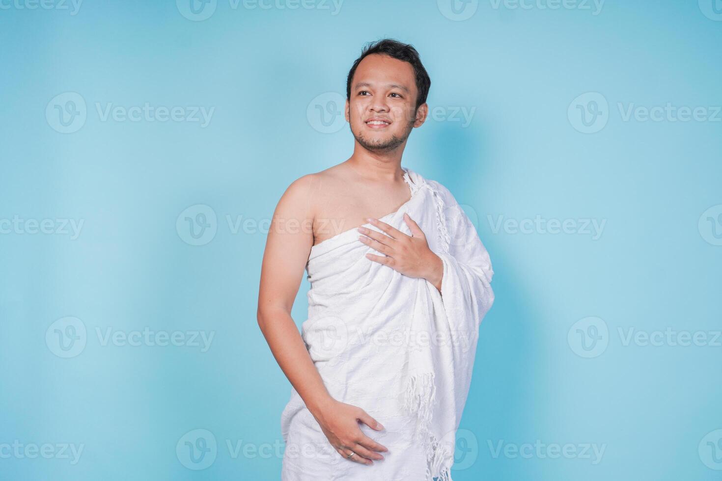 Happy young Asian Muslim man wearing ihram posing hand on chest over isolated blue background. Hajj concept. photo