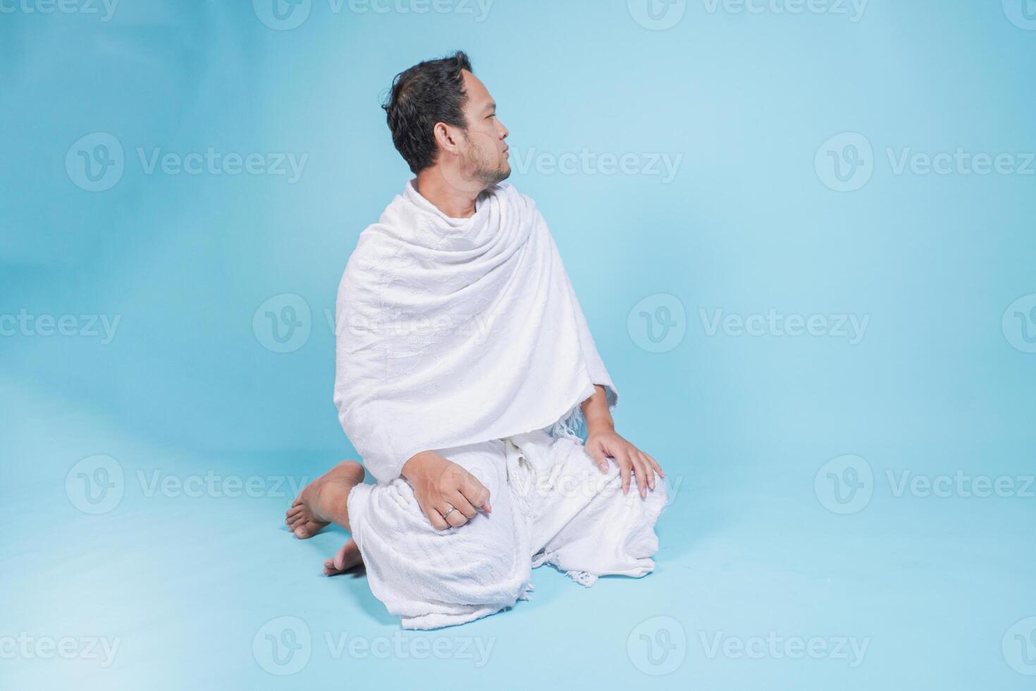 Serious young Asian Muslim man wearing ihram sitting down praying and turning head to the side over isolated blue background. Hajj concept. photo