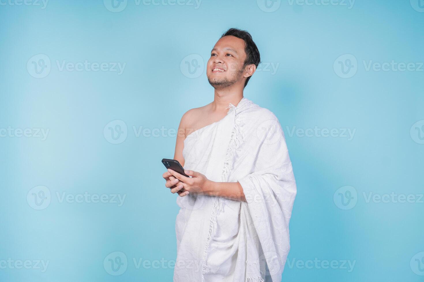Smiling young Asian Muslim man in ihram holding smartphone while lifting his head up over isolated blue background. Hajj concept. photo