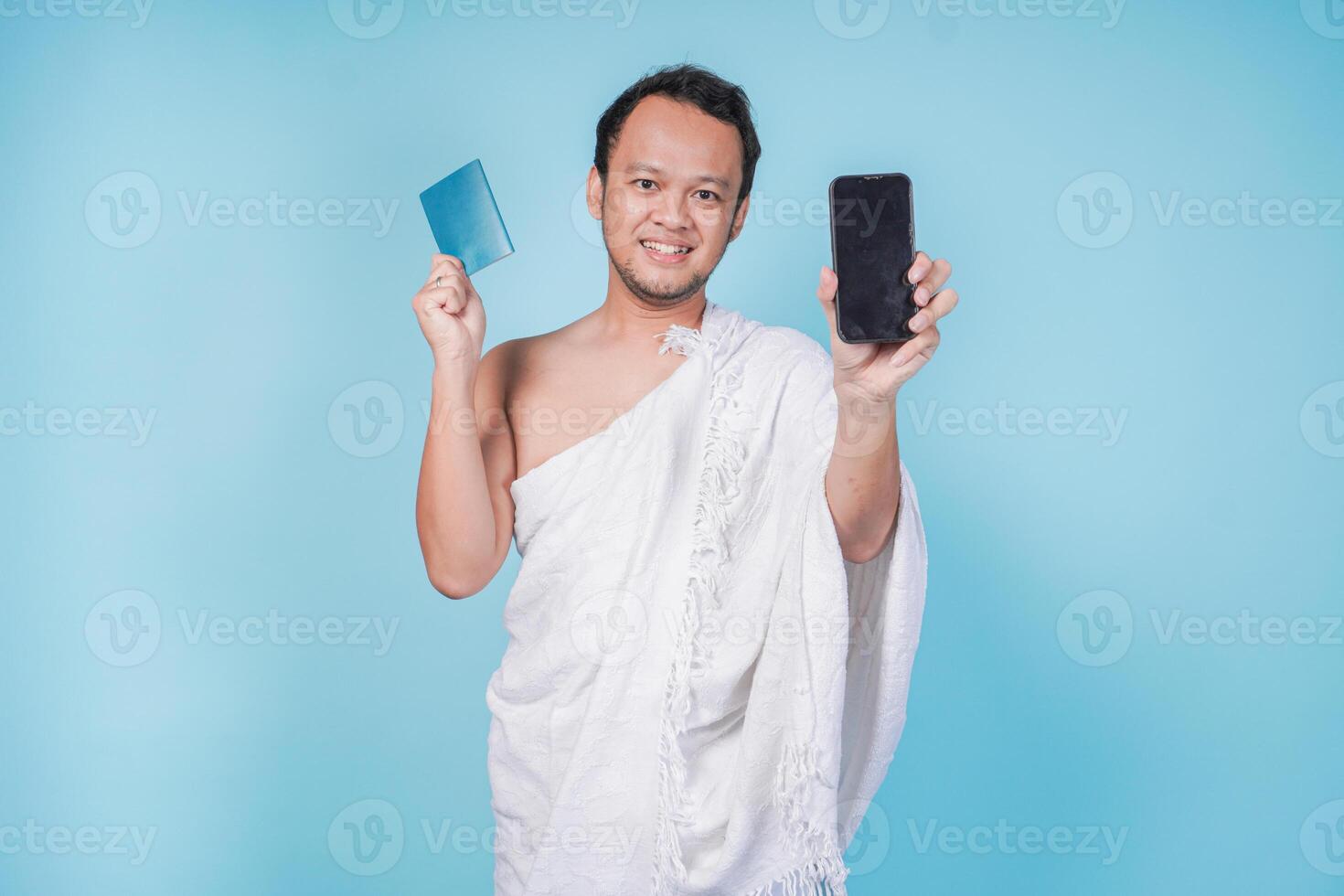 Excited Asian Muslim man in ihram holding passport and smartphone presenting copy space on screen, isolated by blue background. Hajj concept. photo