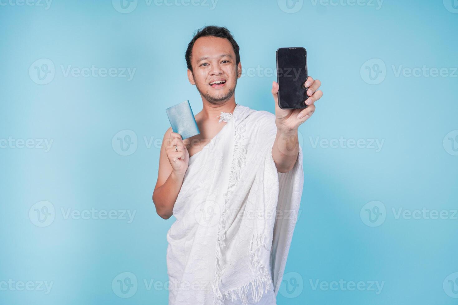 Excited Asian Muslim man in ihram holding passport and smartphone presenting copy space on screen, isolated by blue background. Hajj concept. photo