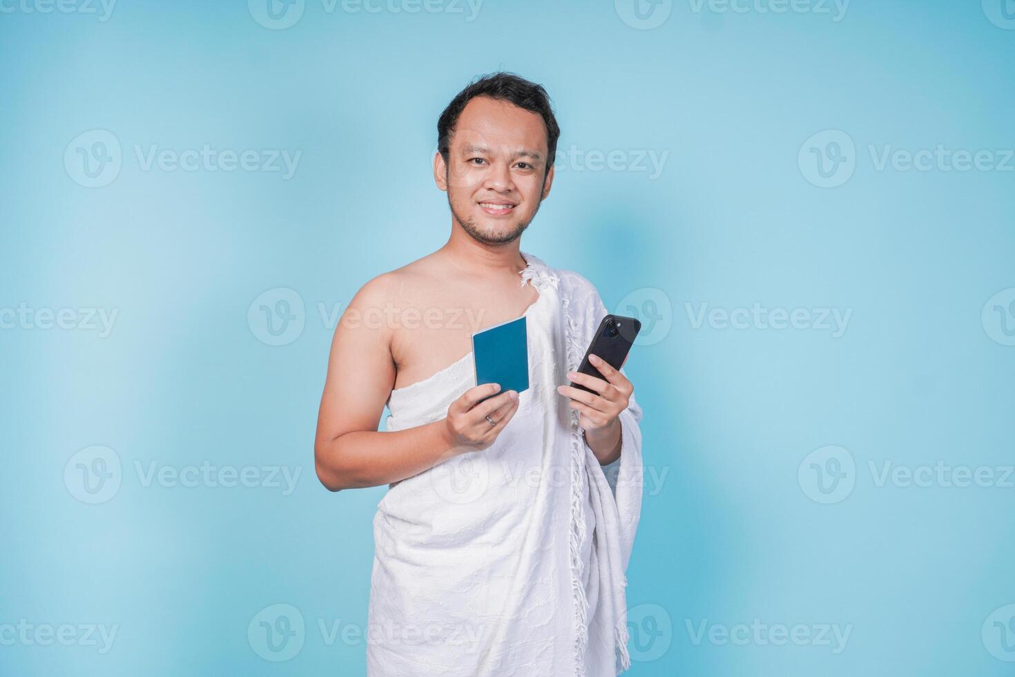 Cheerful Asian Muslim man wearing ihram holding smartphone and passport while smiling, isolated blue background. Hajj concept. photo