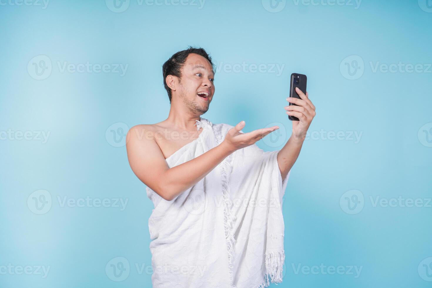 Young Asian Muslim man wearing ihram doing call and pointing to the smartphone he holds, isolated blue background. Hajj concept. photo