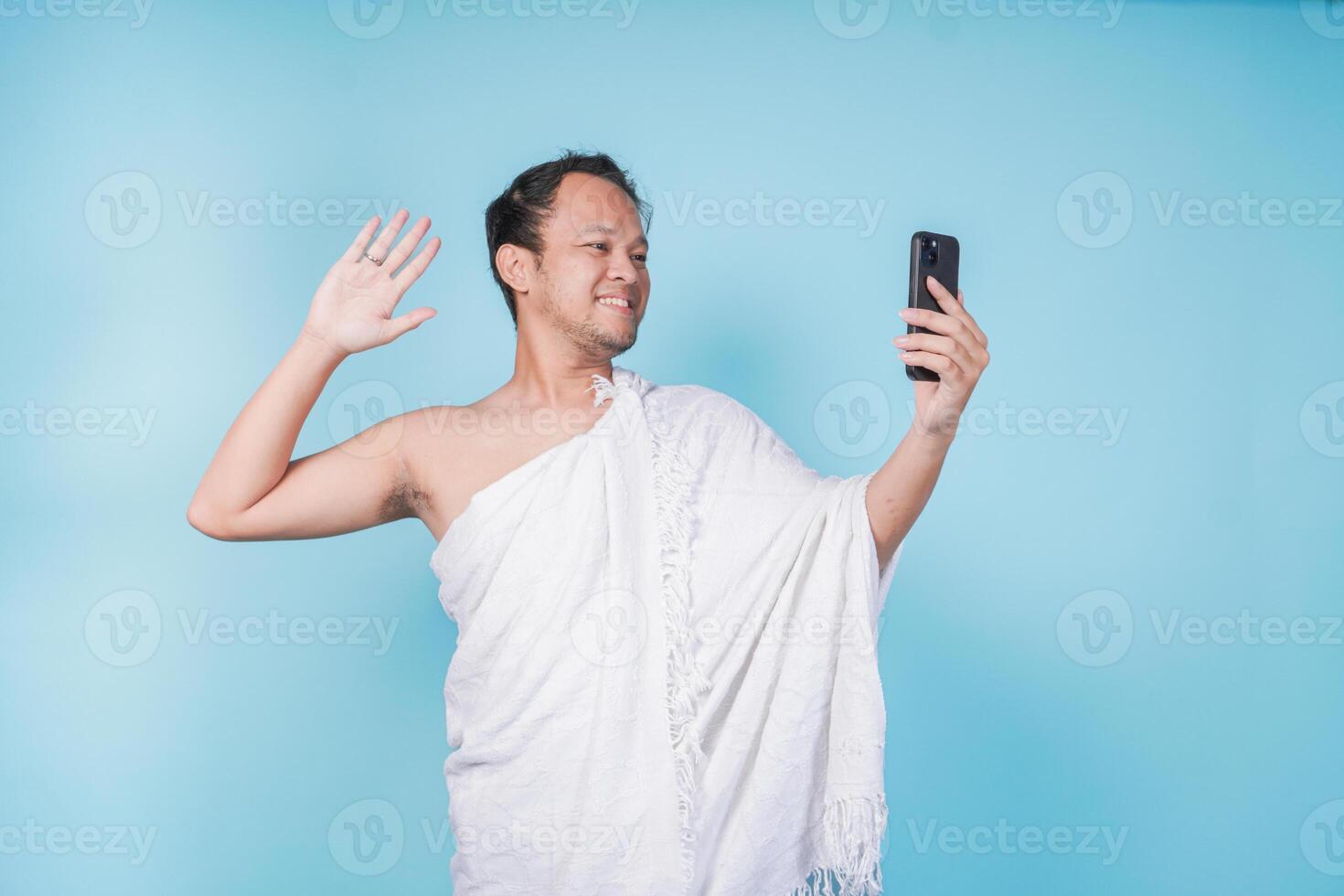 Handsome Asian Muslim man wearing ihram doing call and waving hello gesture, isolated blue background. Hajj concept. photo