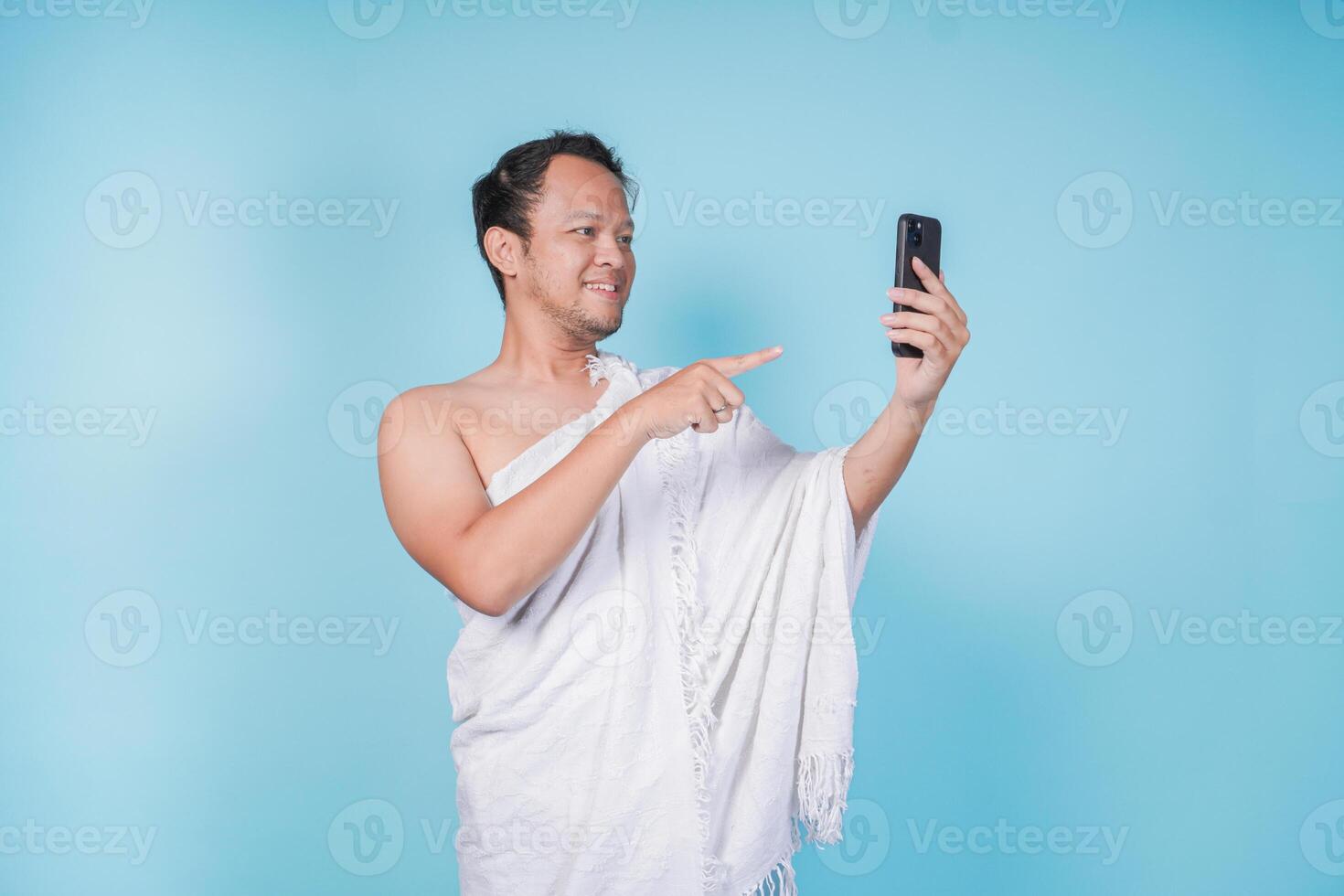 Young Asian Muslim man wearing ihram doing call and pointing to the smartphone he holds, isolated blue background. Hajj concept. photo