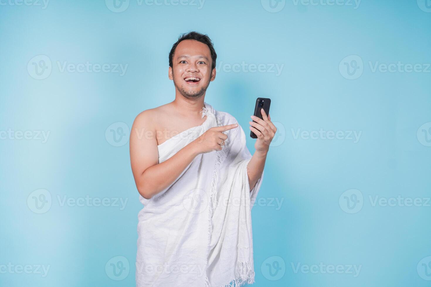 Excited Asian Muslim man wearing ihram pointing to copy space on his smartphone, isolated blue background. Hajj concept. photo