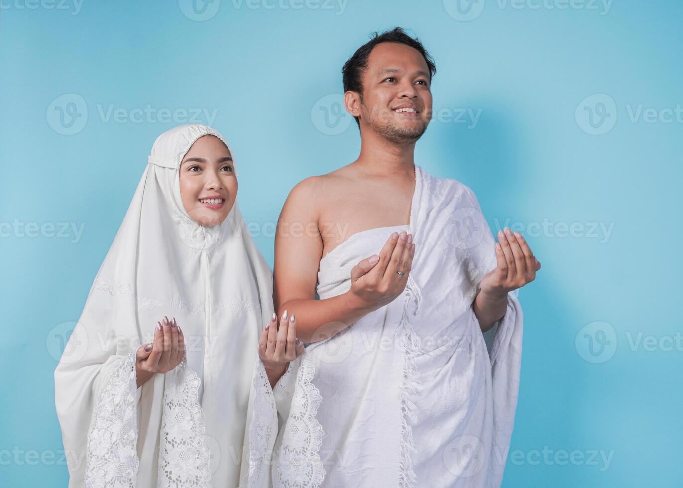 Smiling young Asian Muslim couple wearing ihram and prayer gown with praying hand gesture over isolated by blue background. Hajj concept. photo