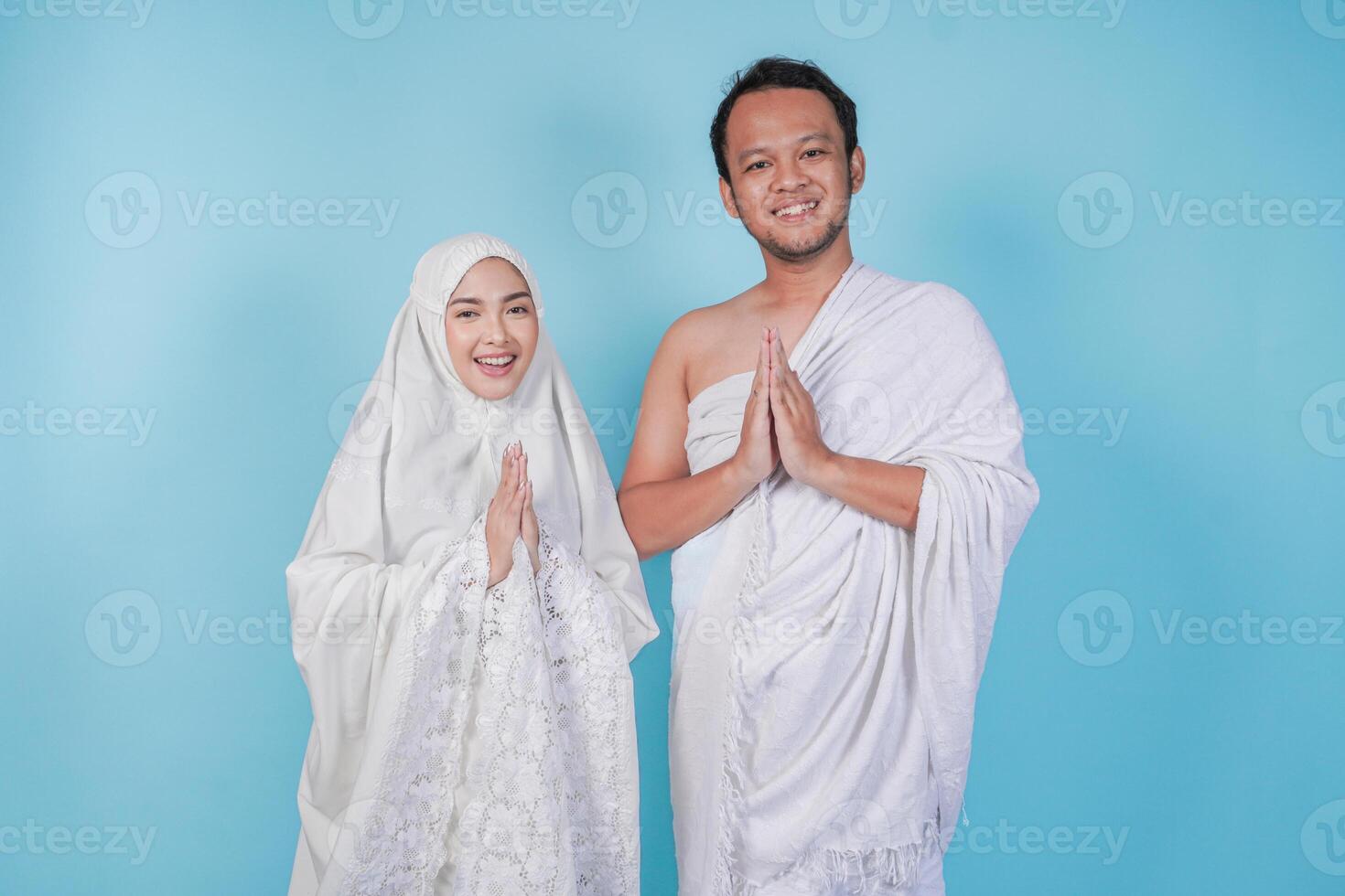 Lovely young Asian Muslim couple in ihram and prayer gown doing traditional greeting gesture, isolated by blue background. Hajj concept. photo