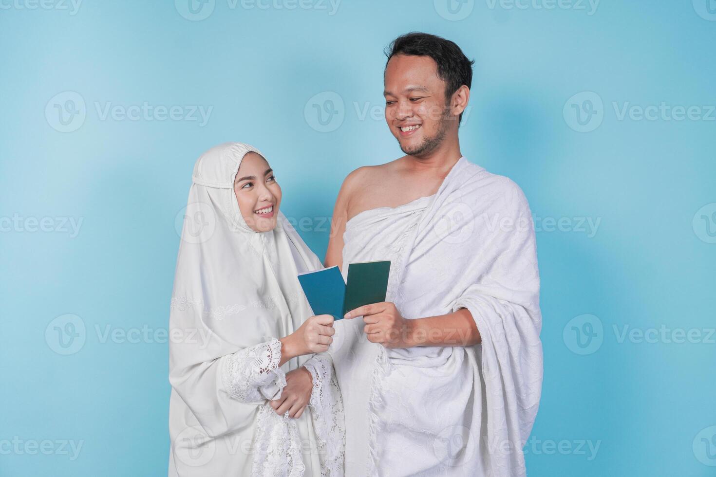 Young Asian Muslim couple in ihram and prayer gown holding a passport while looking at each other, isolated by blue background. Hajj concept. photo