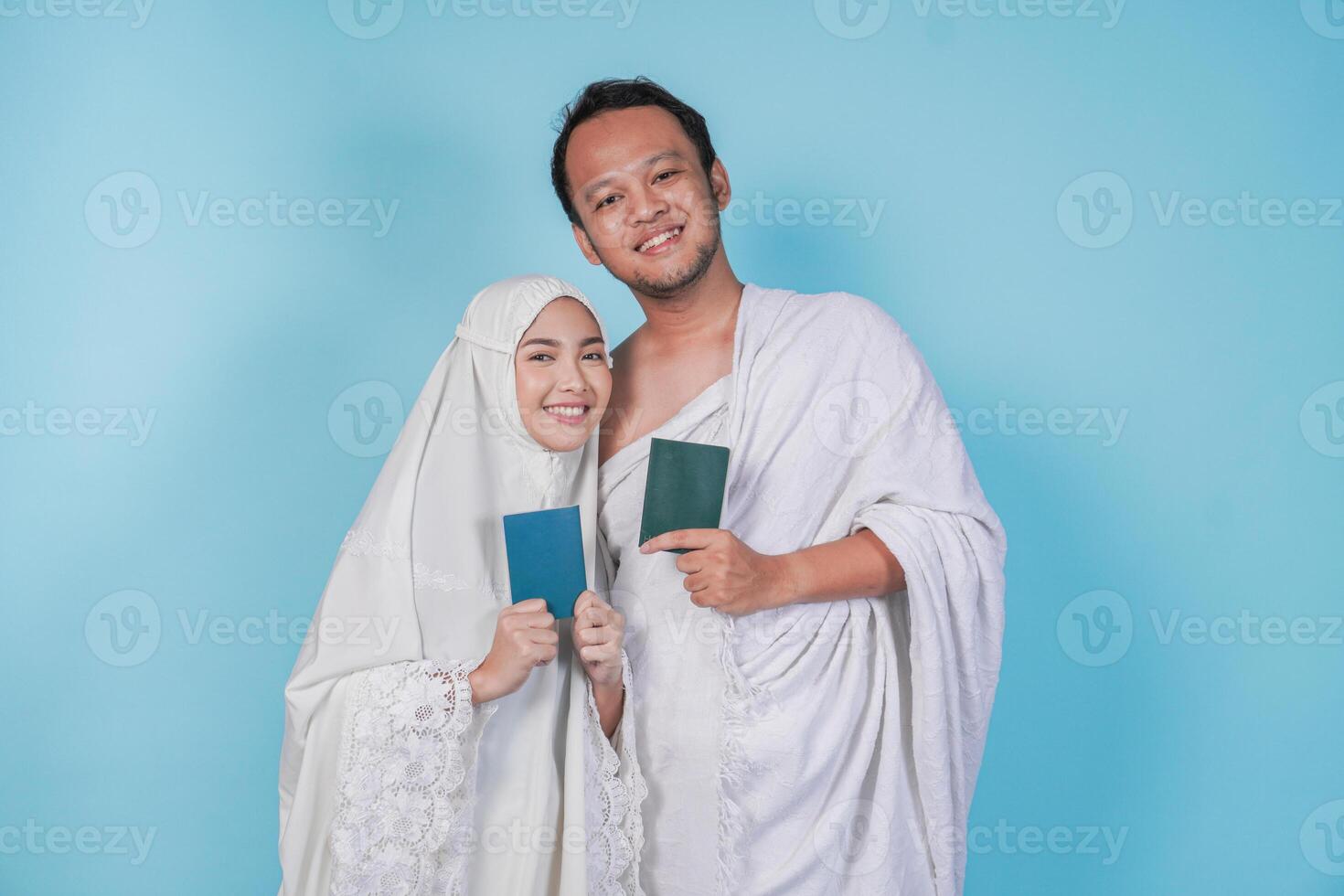 Happy young Asian Muslim couple in ihram and prayer gown posing with a passport over isolated by blue background. Hajj concept. photo