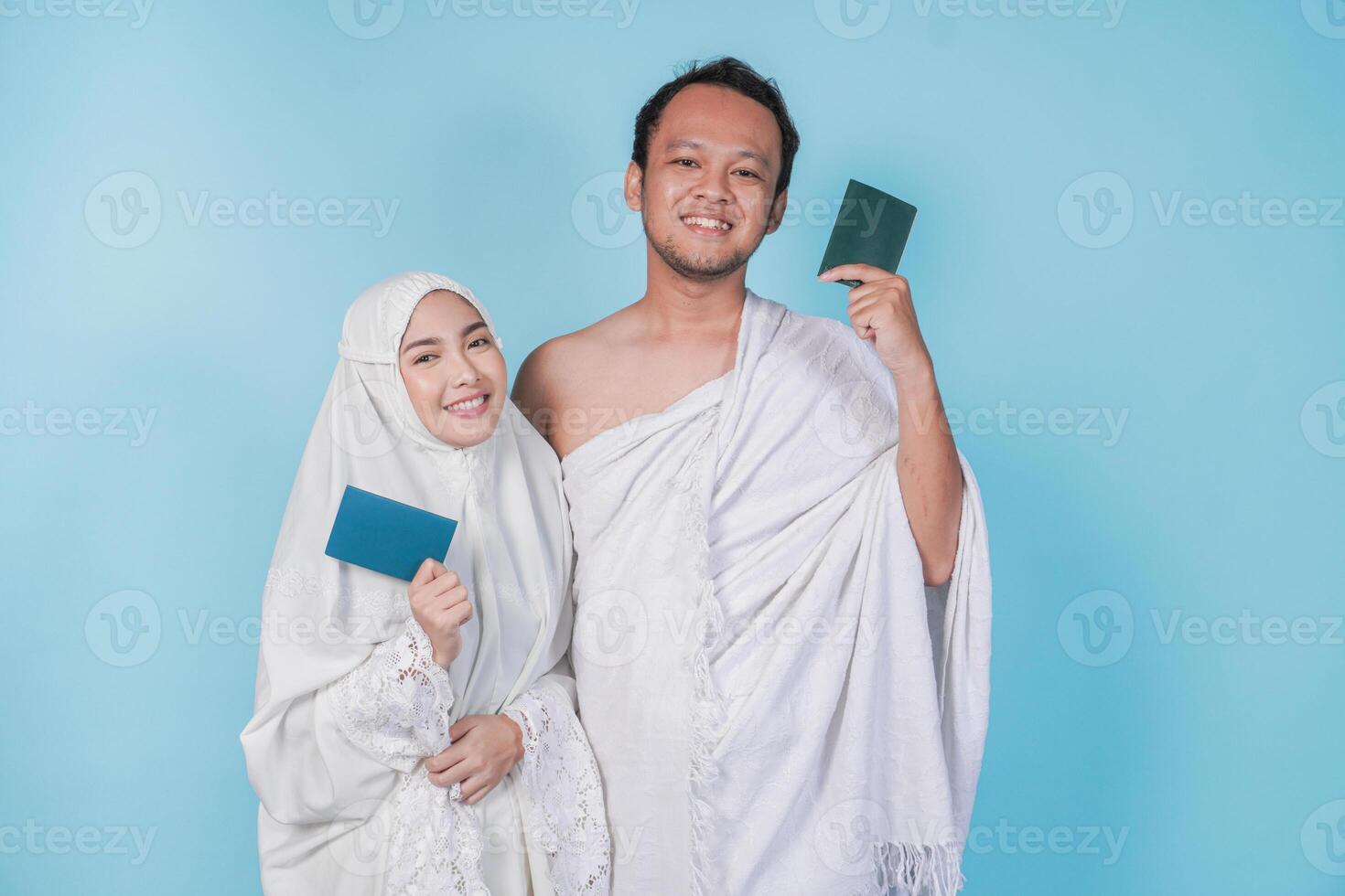 Smiling young Asian Muslim couple wearing ihram and prayer gown holding passport over isolated by blue background. Hajj concept. photo