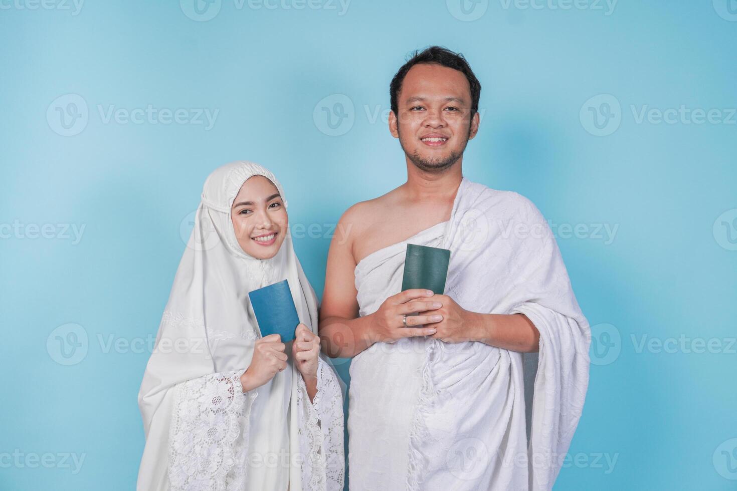 Happy young Asian Muslim couple in ihram and prayer gown posing with a passport over isolated by blue background. Hajj concept. photo