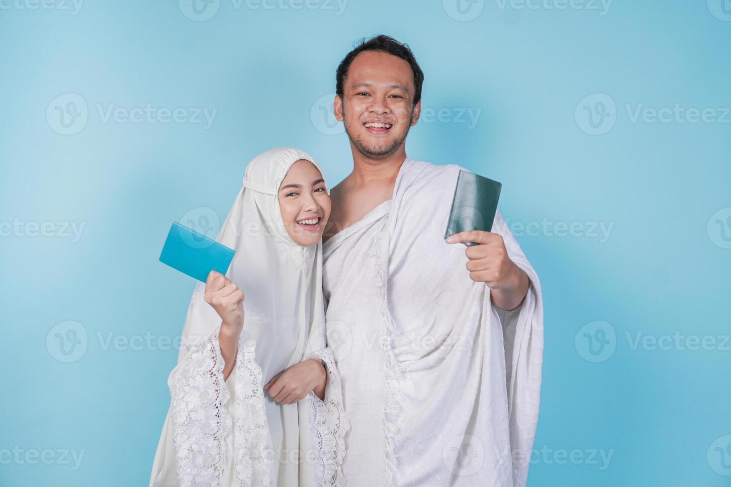 Smiling young Asian Muslim couple wearing ihram and prayer gown holding passport over isolated by blue background. Hajj concept. photo