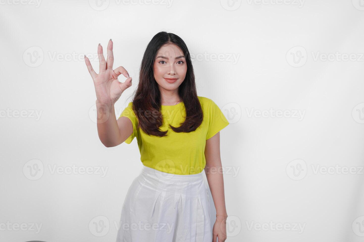 A happy young Asian woman in a yellow shirt and white skirt is giving okay good hand gesture, isolated by white background. photo