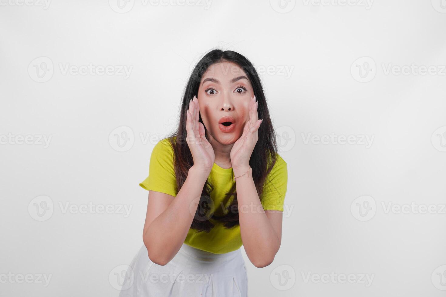Young Asian woman in a yellow shirt is making a whispering gesture with hands on her face, isolated by white background. Concept of surprise or shock. photo