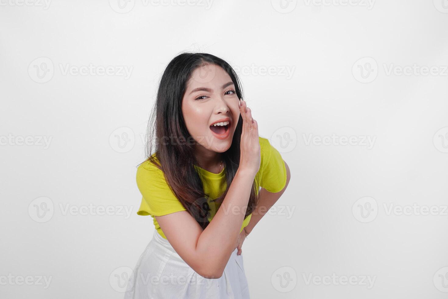 Young Asian woman in a yellow shirt is making a whispering gesture with hands on her face, isolated by white background. Concept of surprise or shock. photo