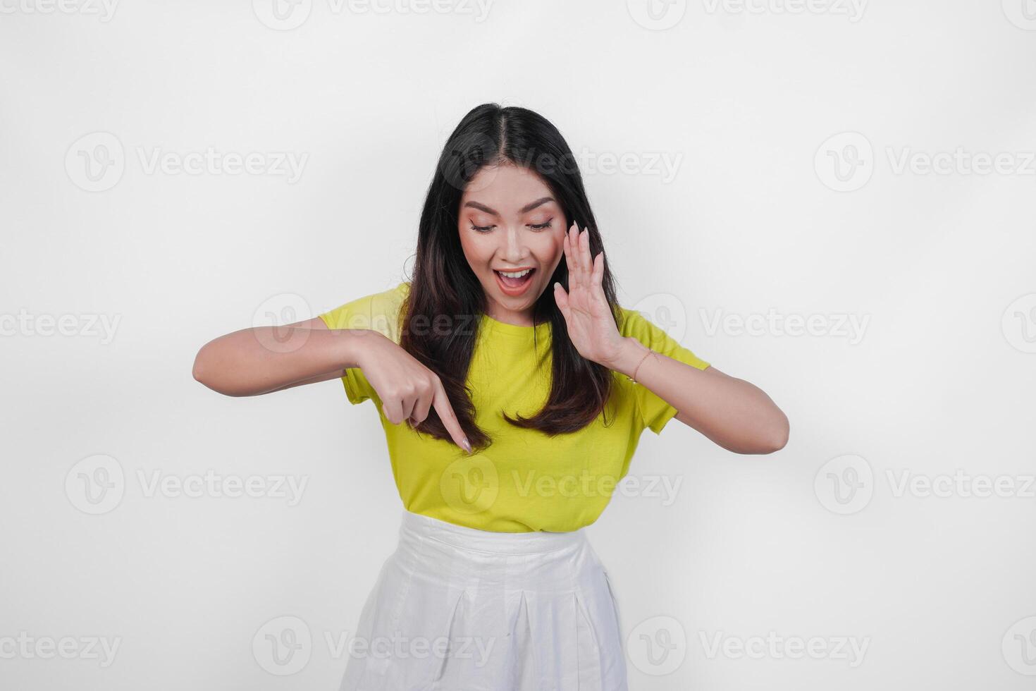 Excited young Asian woman wearing yellow shirt and white skirt making shout gesture while pointing to the copy space downwards, isolated by white background. photo