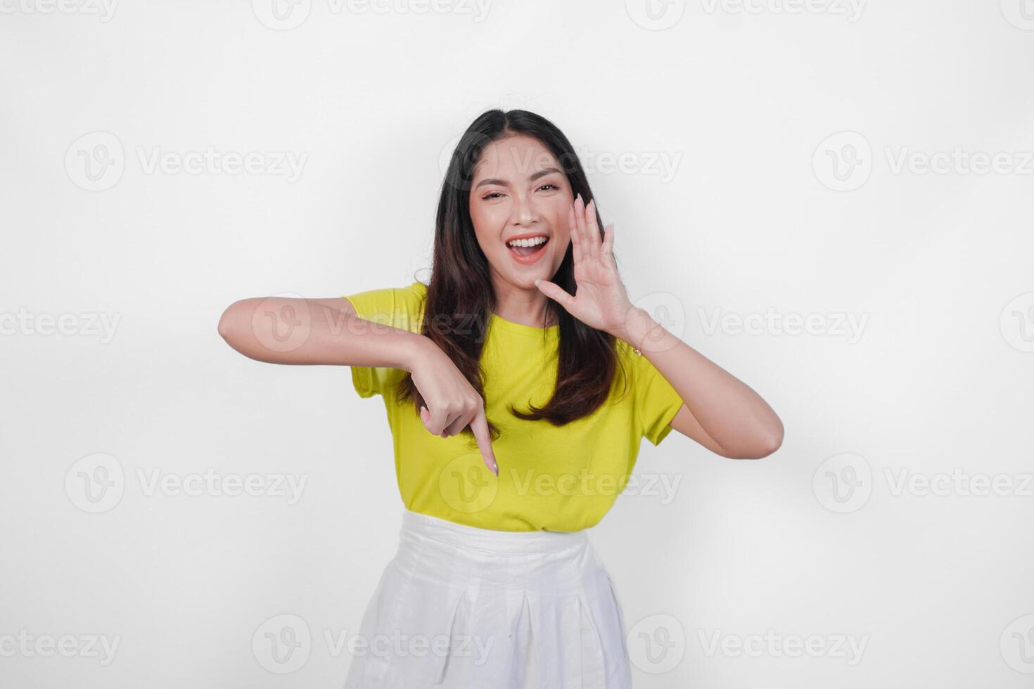 Excited young Asian woman wearing yellow shirt and white skirt making shout gesture while pointing to the copy space downwards, isolated by white background. photo