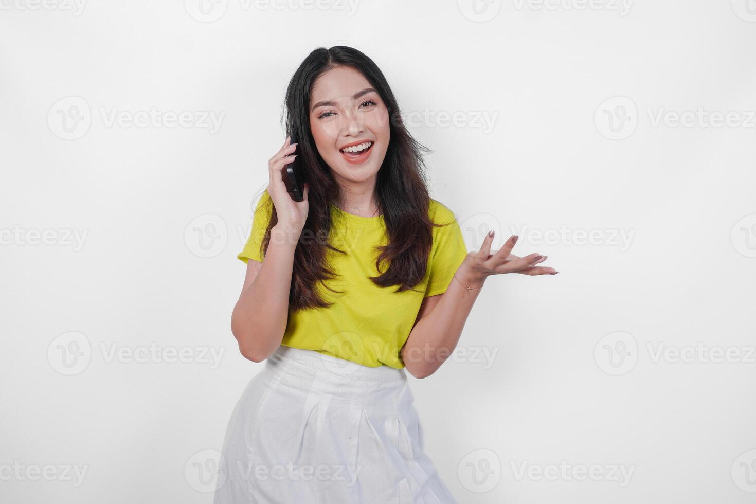 Excited young Asian woman in a yellow shirt is talking on a cell phone while showing copy space beside her, isolated by white background. photo