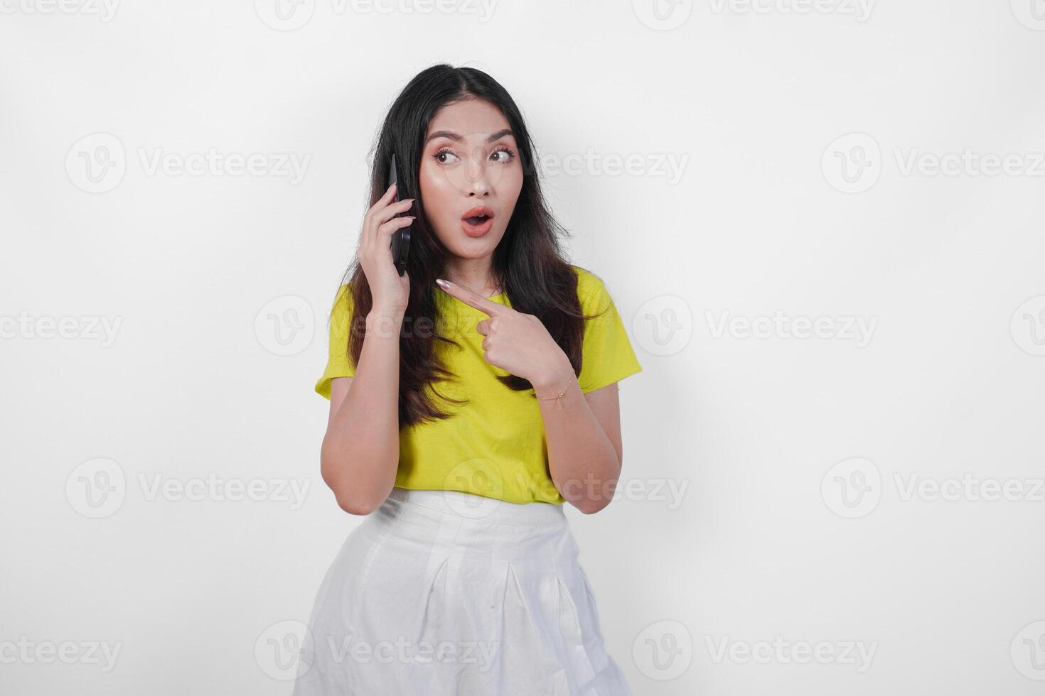 Surprised young Asian woman in a yellow shirt is talking on a cell phone, having a shocking conversation, isolated by white background. photo