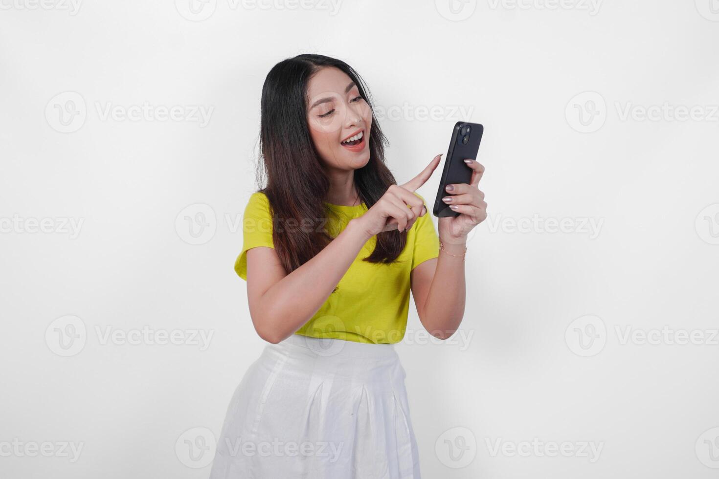 Attractive young Asian woman wearing a yellow shirt and white skirt looking at her smartphone with surprised expression, isolated by white background. photo