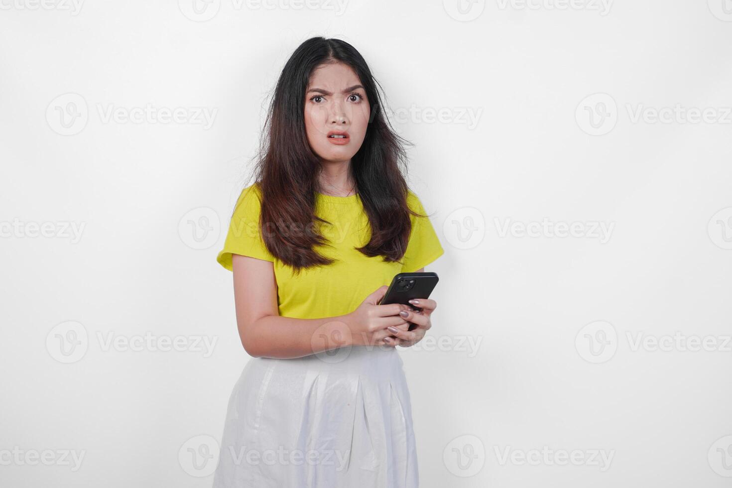 An upset Asian woman in a yellow shirt is holding a black cell phone and appears to be angry. She is looking at the phone with a frown on her face. photo
