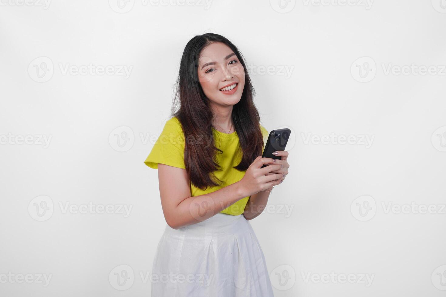 Cheerful Asian woman in a yellow shirt is holding a cell phone, smiling to the camera feeling happy, isolated by white background. photo