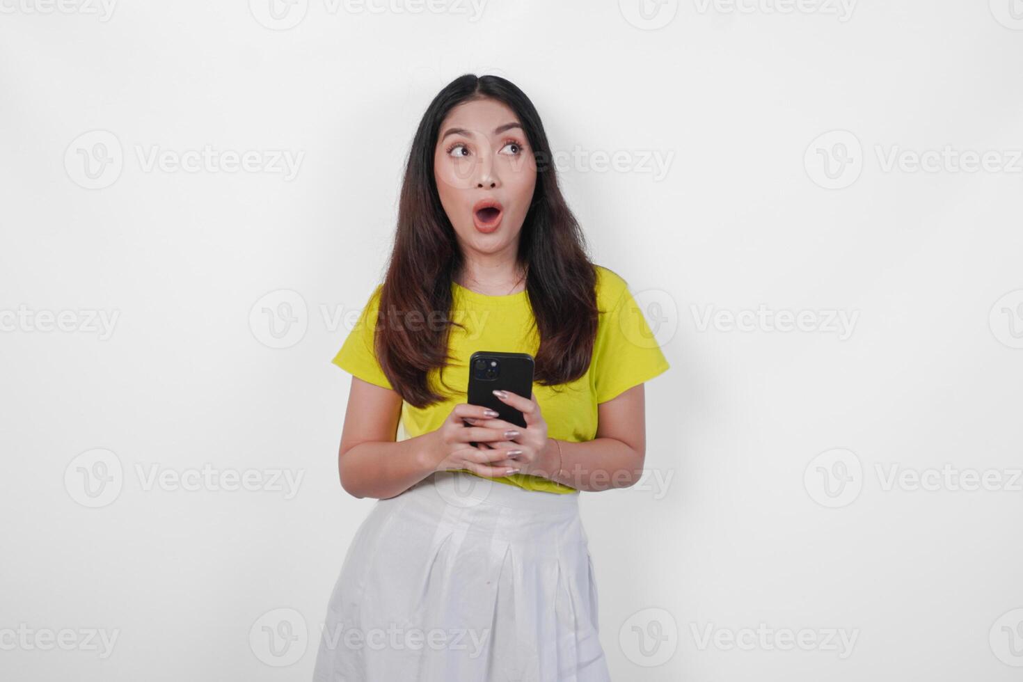Surprised young Asian woman in a yellow shirt pointing at her cell phone over isolated white background. photo