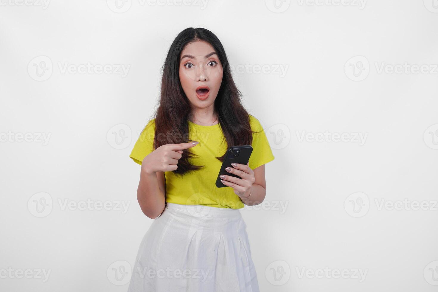 Surprised young Asian woman in a yellow shirt pointing at her cell phone over isolated white background. photo