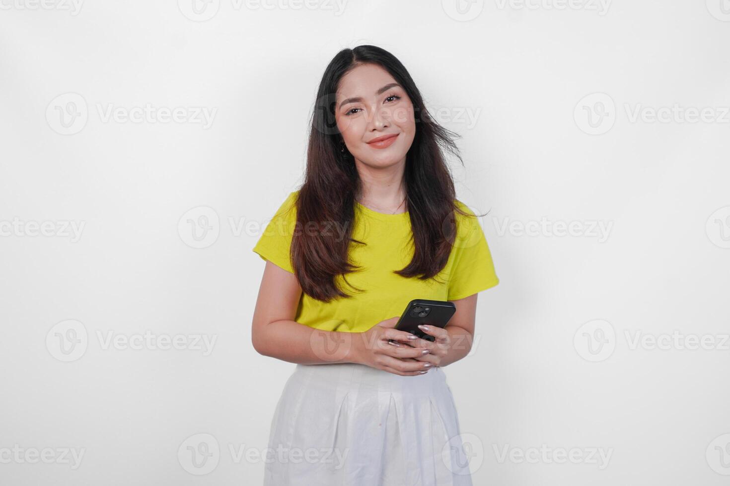 Cheerful Asian woman in a yellow shirt is holding a cell phone, smiling to the camera feeling happy, isolated by white background. photo