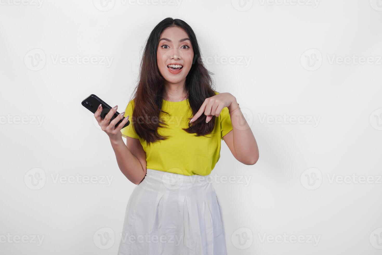 Smiling young woman is holding a cell phone and pointing to the copy space downwards over isolated white background. photo