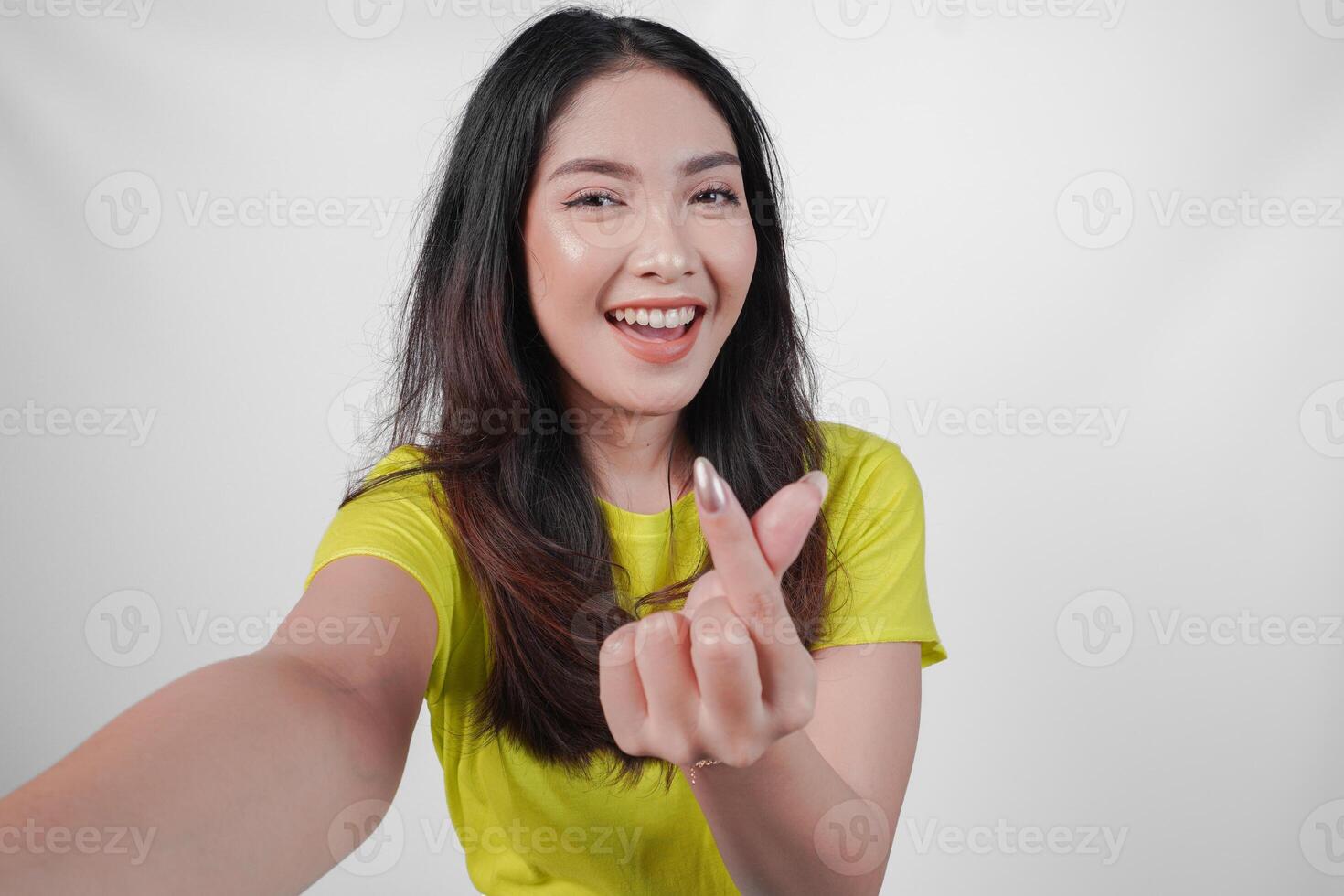 Lovely young Asian woman in a yellow shirt is smiling and showing finger heart gesture to the camera, isolated by white background. photo