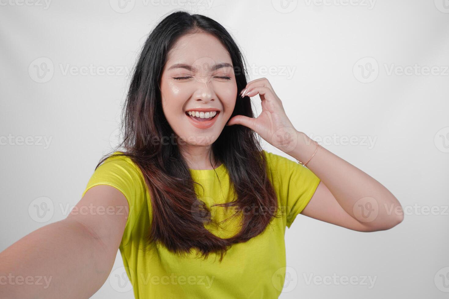 Cute young Asian woman in a yellow shirt is taking a selfie while making a heart gesture on her cheek, isolated over white background. photo