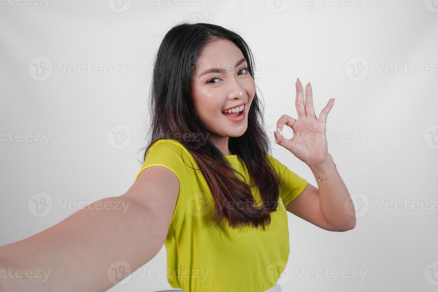 Attractive young Asian woman in a yellow shirt is taking a selfie and giving okay sign. Concept of happiness and positivity, as the woman is smiling and expressing approval. photo